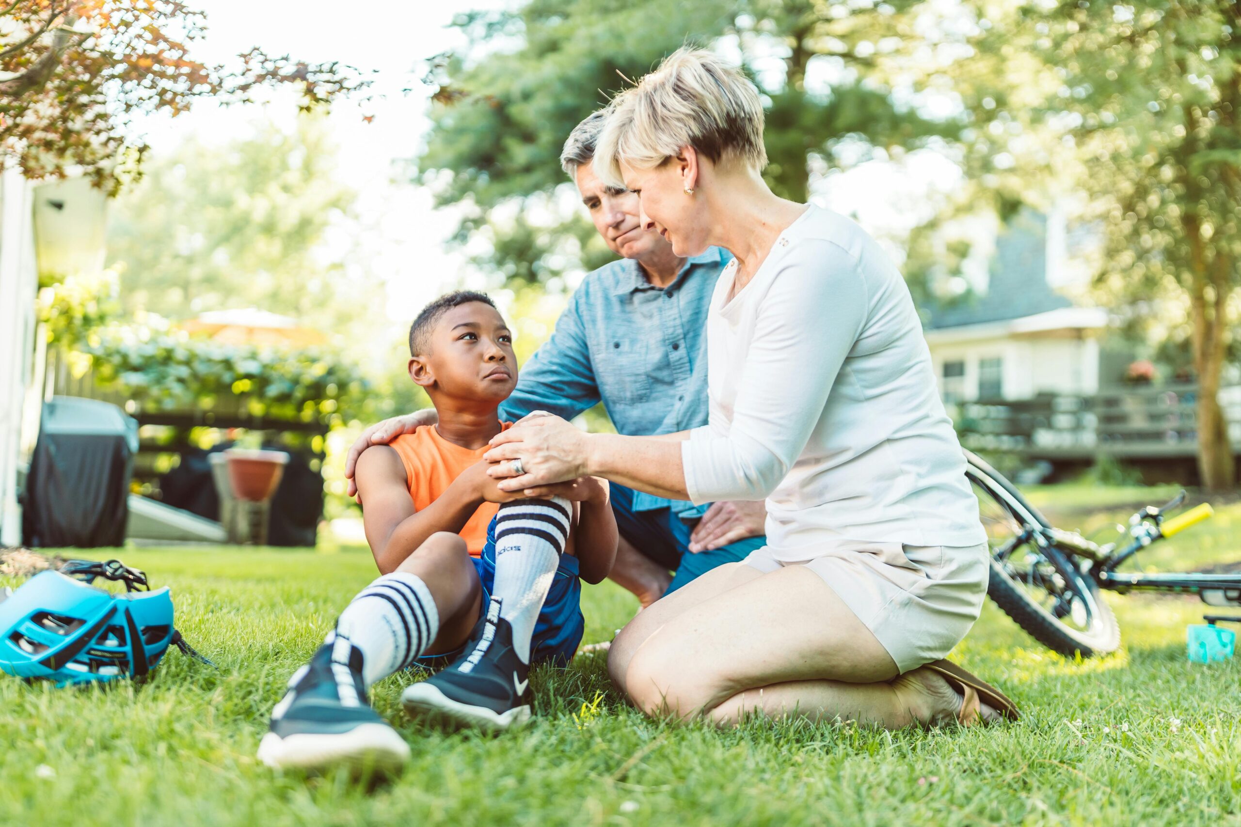 Parents comforting their injured son after a biking accident in a sunny backyard.