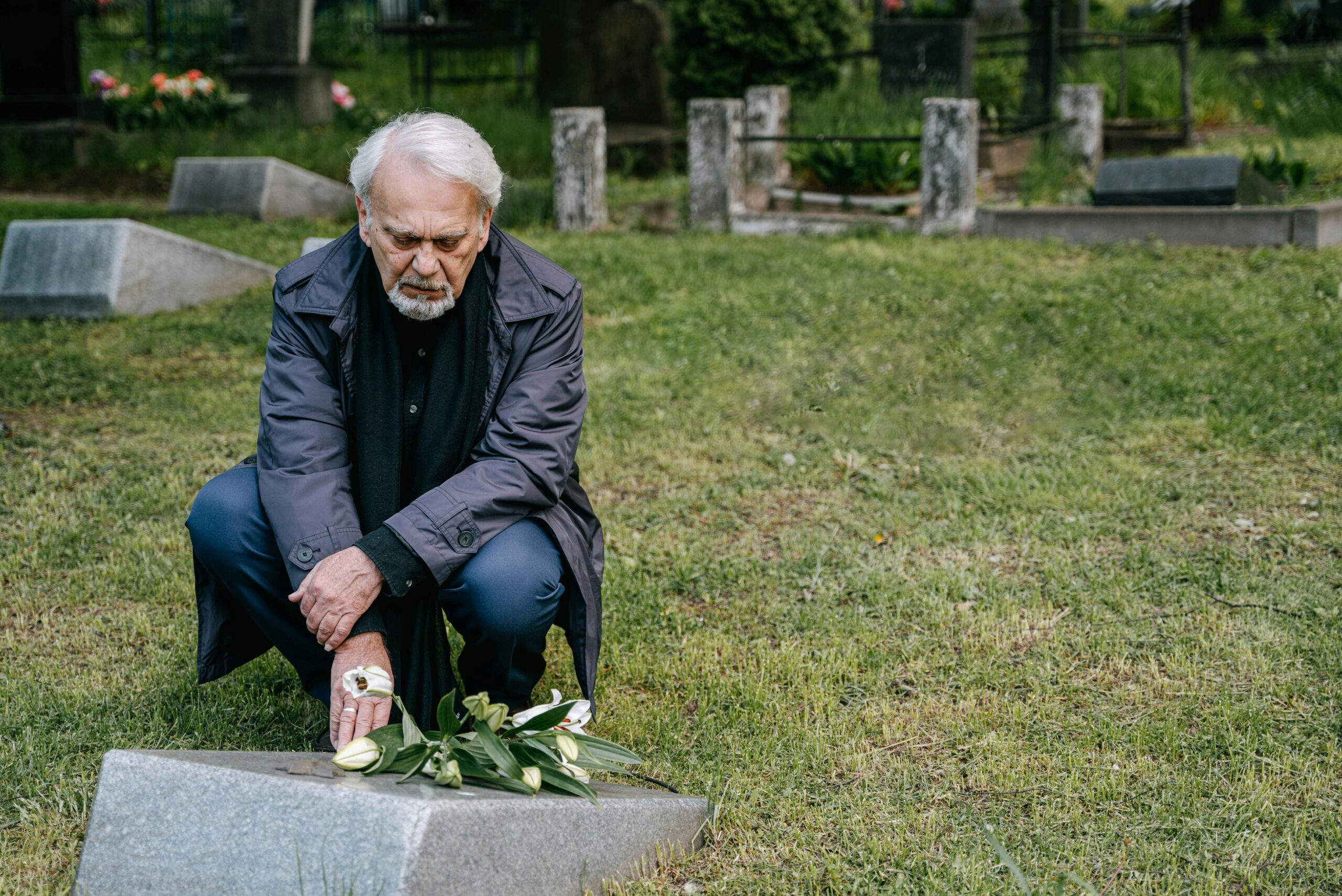 Elderly man kneeling in grief at a cemetery with flowers placed on a headstone.
